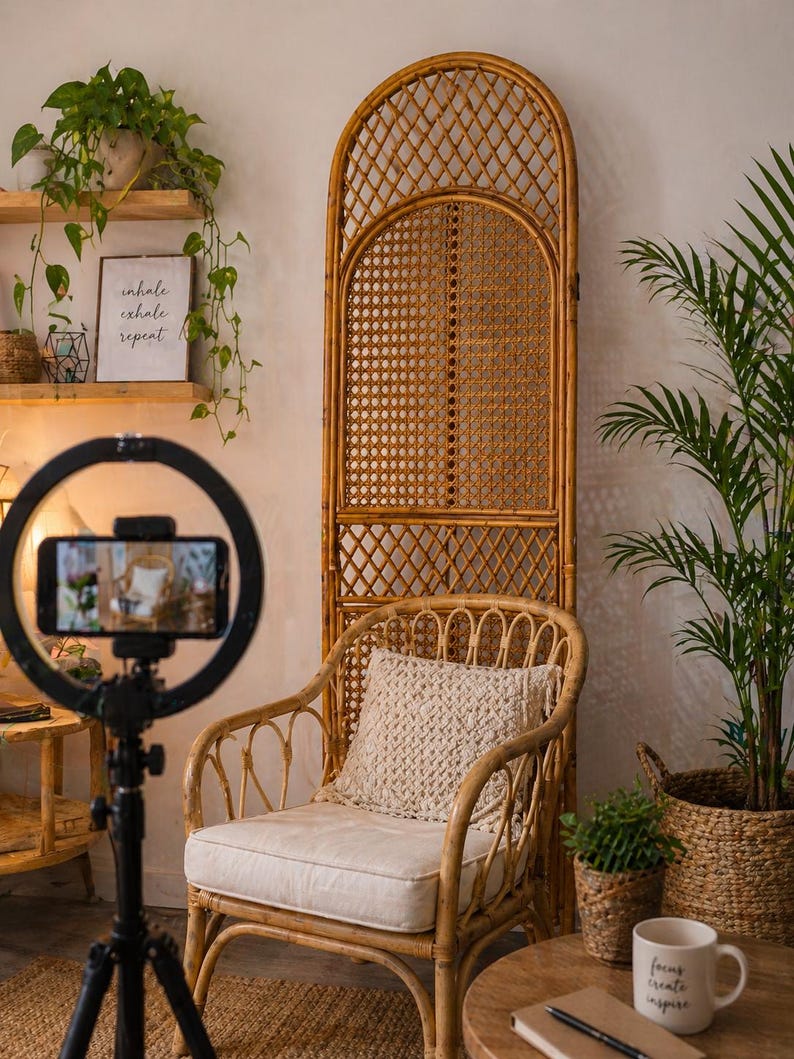 May include: A cozy interior scene featuring a rattan chair with a cream cushion and a macrame pillow. A tall, arched rattan screen stands behind the chair. A ring light with a phone is in the foreground. Plants and a mug with text add to the warm, inviting atmosphere.