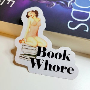 May include: A white sticker with a pin-up style illustration of a woman sitting on a stack of books. The text "Book Whore" is printed in black below the illustration.