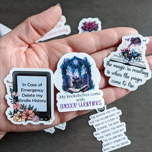 May include: Four book-themed stickers with colorful designs and text. The stickers feature a Kindle e-reader, a bookshelf with a gothic window, a book with flowers and a butterfly, and a book with the text "The magic in reading is when the pages come to life."