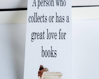 May include: A white bookmark with a black and white illustration of a person holding a stack of books. The bookmark has the text "Bib-li-o-phile" and "A person who collects or has a great love for books".