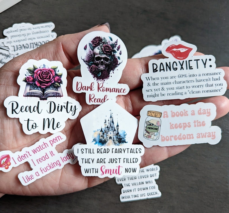 May include: A collection of colorful stickers with various book-themed quotes and illustrations. The stickers feature a skull with roses, a castle, a book with roses, a jar of books, and a quote about reading dirty books. The stickers are printed on white backgrounds.