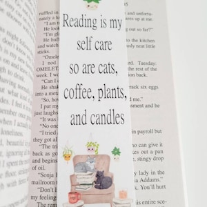 Könnte beinhalten: Ein weißes Lesezeichen mit einer handgezeichneten Illustration von zwei Katzen, die auf einem Stapel Bücher in einem gemütlichen Sessel sitzen. Das Lesezeichen zeigt den Text "Reading is my self care, so are cats, coffee, plants, and candles".