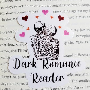 Könnte beinhalten: Ein weißer Aufkleber mit schwarzem Text, der "Dark Romance Reader" lautet. Der Aufkleber zeigt eine Schwarzweißillustration von zwei Skeletten, die sich umarmen. Die Skelette sind von rosa und roten Herzen umgeben.