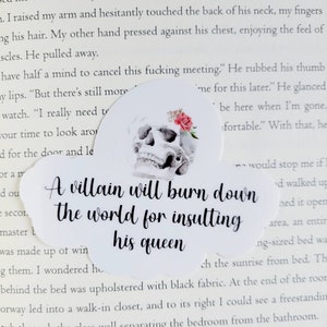 May include: A white sticker with a skull illustration and a floral accent. The sticker has the text "A villain will burn down the world for insulting his queen" in a handwritten style font.