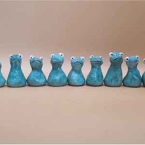 May include: A row of nine teal ceramic frog ornaments with white eyes. Each frog has a different facial expression, from cheerful to glum. The ornaments are displayed in a line, highlighting their diverse designs.