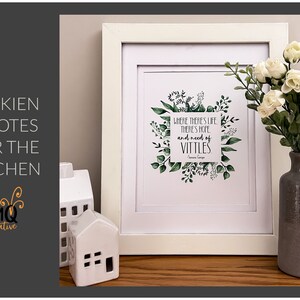 May include: A white framed print with a green leafy border and a quote from J.R.R. Tolkien: "Where there's life there's hope and need of vittals." The print is sitting on a wooden surface with a white ceramic house and a gray vase with white flowers.
