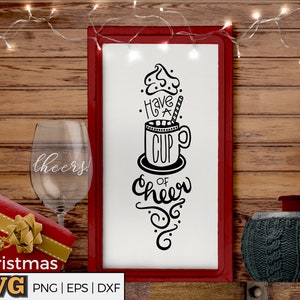 Könnte beinhalten: Ein gerahmtes Bild mit dem Text "Have a Cup of Cheer" und einer Tasse mit Sahnehaube. Ein Weinglas mit "cheers", eine rote Geschenkbox und eine Tasse mit Strickmuster sind ebenfalls im Bild zu sehen.