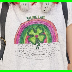 May include: A white t-shirt with a rainbow and a green four-leaf clover drawn on it. The text "Shine Like A Shamrock" is written below the drawing.