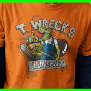 Puede incluir: Camiseta naranja con un dinosaurio de dibujos animados que lleva un mono y sostiene un martillo. El dinosaurio está de pie sobre una excavadora y el texto "T. Wreck's Demo Squad" está impreso en la camiseta.