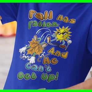 Puede incluir: Una camiseta azul con una ilustración de dibujos animados de un sol pateando una hoja. El texto en la camiseta dice "Fall has fallen, and he can't get up!"