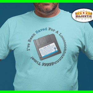 Peut inclure: Un t-shirt bleu clair avec une illustration en noir et blanc d'une disquette avec le texte "I've Been Saved For A Looooong Time!" et le verset biblique "Actes 4:12".