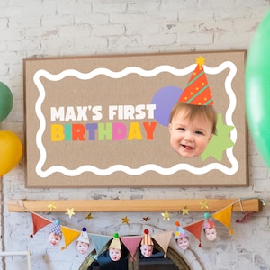 May include: A framed sign with the text "MAX'S FIRST BIRTHDAY" in colorful letters, featuring a baby's face wearing a party hat. Balloons in red, yellow, green, and blue are on the left. A banner with party hats and faces hangs below.