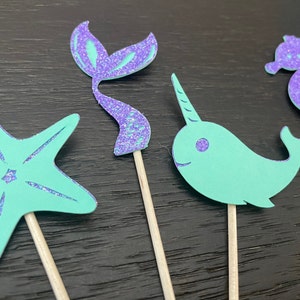 May include: Four whimsical cupcake toppers featuring a starfish, mermaid tail, narwhal, and seahorse. Each figure is a combination of teal and purple glitter, mounted on a wooden stick. These decorative toppers are perfect for a mermaid or ocean-themed party.