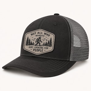 May include: A black baseball cap with a grey mesh back and a beige patch on the front. The patch features a black graphic of a Bigfoot silhouette, mountains, and pine trees with the text "NOT ALL WHO WANDER ARE LOOKING FOR PEOPLE".
