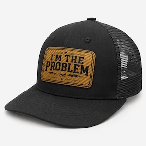 May include: A black trucker cap featuring a rectangular tan patch with a carbon fibre texture. The patch displays the black text I'M THE PROBLEM, a small skull icon, and two lightning bolt graphics. The cap has a solid front and mesh back.