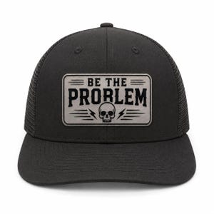 Op de afbeelding: Een zwarte truckerpet met een rechthoekige grijze patch aan de voorzijde. De patch toont de zwarte tekst BE THE PROBLEM boven een afbeelding van een schedel geflankeerd door twee bliksemschichten en horizontale lijnen op een lichtgrijze achtergrond.