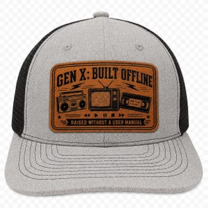 May include: A grey and black trucker hat featuring a rectangular brown patch on the front. The patch displays black graphics of a boombox, a television, and a VHS tape, with the text GEN X: BUILT OFFLINE and RAISED WITHOUT A USER MANUAL.