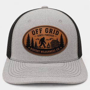 Könnte beinhalten: Eine graue und schwarze Trucker-Cap mit einem braunen, ovalen Aufnäher auf der Vorderseite. Der Aufnäher zeigt eine schwarze Grafik mit einer Bigfoot-Silhouette, einer Hütte, Bäumen und Bergen sowie den Text OFF GRID SINCE FOREVER BIGFOOT WILDERNESS CO.