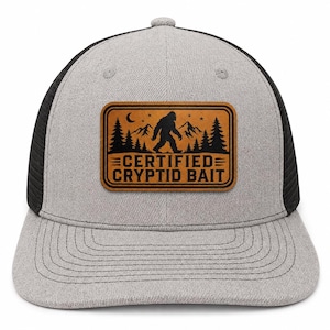Può includere: Un cappellino trucker grigio e nero con una toppa rettangolare in pelle color cuoio sul davanti. La toppa presenta una grafica nera con la sagoma di un Bigfoot, montagne, alberi, una luna crescente e stelle, con il testo CERTIFIED CRYPTID BAIT.