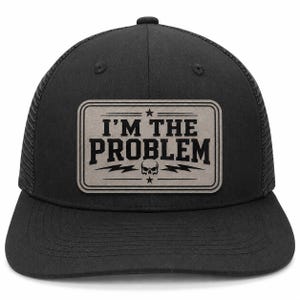May include: A black trucker hat featuring a rectangular grey patch on the front. The patch displays the text I'M THE PROBLEM in bold black letters, accompanied by a small skull graphic, two lightning bolts, and two stars within a double-lined border.