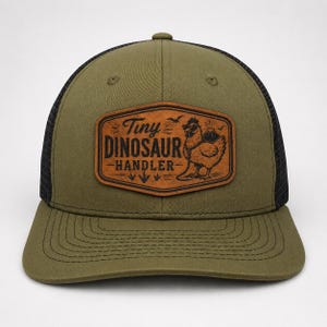 May include: An olive green baseball cap with a black mesh back and a brown leather-like patch on the front. The patch features a graphic of a chicken wearing sunglasses with the text Tiny DINOSAUR HANDLER and a small volcano illustration.