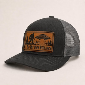 May include: A black trucker hat featuring a rectangular brown leather patch on the front. The patch displays a graphic of Bigfoot and a UFO above a forest, with the text I DO MY OWN RESEARCH printed in a bold, dark font below the illustration.