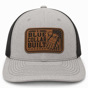 May include: A heather grey baseball cap with black mesh back panels and a brown rectangular leather patch on the front. The patch features the text BLUE COLLAR BUILT, an illustration of work gloves, and a stylised American flag design.