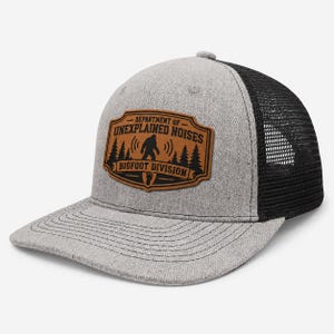 Può includere: Un cappellino da baseball grigio mélange con retro in rete nera e visiera piatta. Una toppa color cuoio sul davanti presenta una grafica nera con la sagoma di Bigfoot, alberi di pino e il testo: DEPARTMENT OF UNEXPLAINED NOISES BIGFOOT DIVISION.