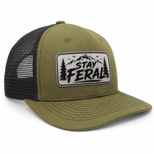 May include: An olive green baseball cap with a black mesh back and a flat brim. A rectangular patch on the front features a black graphic of mountains, pine trees, and birds with the text STAY FERAL printed in a bold, stylized black font.