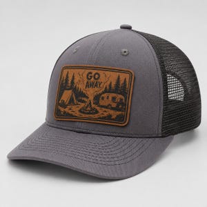 May include: A charcoal grey trucker hat featuring a rectangular brown leather patch on the front. The patch displays a black engraved illustration of a tent, a campfire, and a camper trailer with the text GO AWAY centred above the campfire.