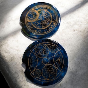 May include: Two round car coasters with a blue galaxy design and gold geometric patterns. Each coaster has a small notch for easy removal. The coasters are made of a glossy material and are placed on a light gray surface.