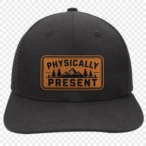 May include: A black trucker hat featuring a rectangular tan patch on the front. The patch displays the text PHYSICALLY PRESENT in black capital letters, with a graphic of a mountain range and pine trees centered between the two lines of text.