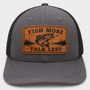 May include: A charcoal gray baseball cap with a black mesh back and a rectangular tan leather patch on the front. The patch features a black graphic of a fish jumping out of water, pine trees, and the text FISH MORE TALK LESS in black.