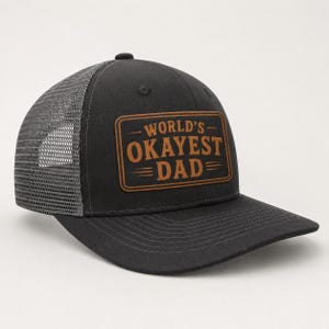 May include: A black trucker hat with a grey mesh back and a rectangular brown leather patch on the front. The patch features the text "WORLD'S OKAYEST DAD" in a bold, dark brown font, framed by horizontal lines above and below the lettering.