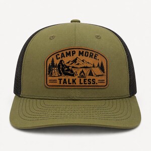 May include: An olive green baseball cap with a black mesh back and a brown leather-like patch on the front. The patch features a black graphic of a creature, a tent, a campfire, and mountains with the text CAMP MORE. TALK LESS. in black.