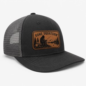 Pode incluir: Um boné trucker preto com rede cinza na parte de trás e um emblema retangular de couro marrom na frente. O emblema apresenta um gráfico preto de uma criatura pescando perto de um lago com pinheiros, montanhas e o texto GONE SQUATCHIN'.