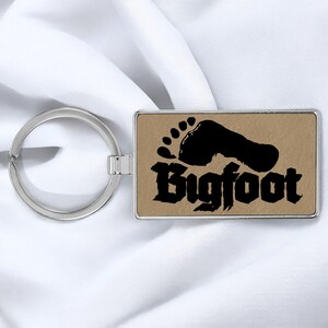 May include: A silver key chain with a rectangular tag. The tag has a brown background with a black footprint and the word "Bigfoot" in black stylized lettering.