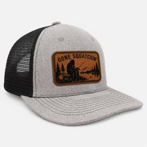 May include: A heather gray and black trucker hat featuring a rectangular brown leather patch on the front. The patch displays a black graphic of a creature fishing by a lake with pine trees and mountains, and the text GONE SQUATCHIN'.