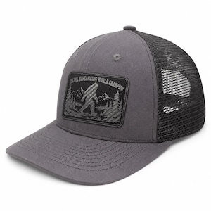 Può includere: Un cappellino trucker grigio antracite con retro in rete nera e una toppa rettangolare sul davanti. La toppa presenta una grafica nera e grigia di una silhouette di Bigfoot in una foresta con montagne e il testo SOCIAL DISTANCING WORLD CHAMPION.