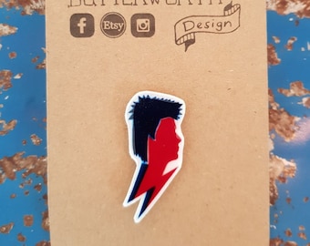Puede incluir: Un pin de esmalte blanco con una silueta roja y azul de la cara de David Bowie y un rayo. El pin está en una tarjeta marrón con el texto "Sophie Butterworth Design" y los iconos de las redes sociales.