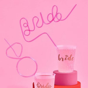 May include: Two clear plastic cups with pink and gold lettering. One cup says "bride" and the other says "bride's babes". A pink bendy straw is shaped to spell "bride" and is in the background.