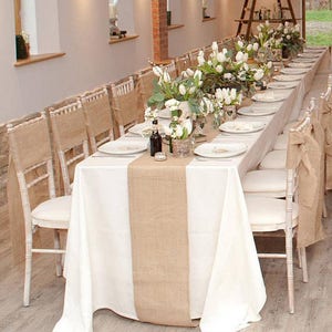 May include: A white tablecloth with a burlap runner down the center. The table is set with white plates, silverware, and glasses. There are white flowers in vases down the center of the table. The chairs are white with burlap sashes.