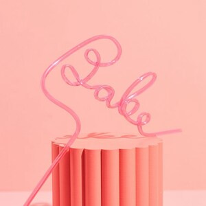 May include: Pink plastic straw shaped to spell the word "babe" on a pink cylindrical pedestal.