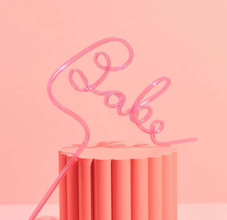 May include: Pink plastic straw shaped to spell the word "babe" on a pink cylindrical pedestal.