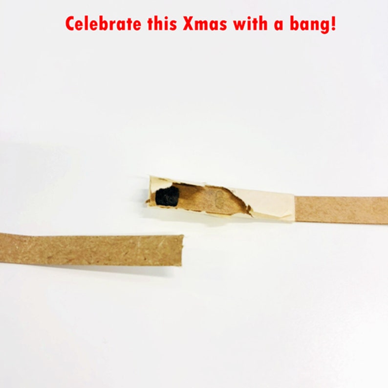 May include: A close-up of a Christmas cracker, with a torn section revealing a small black object. The cracker is made of brown cardboard and has a white paper section. The text "Celebrate this Xmas with a bang!" is in red.
