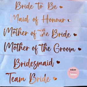 Peut inclure: Six rubans de satin blanc avec des lettres dor&eacute;es roses. Les rubans indiquent "Bride To Be", "Maid of Honour", "Mother of the Bride", "Mother of the Groom", "Bridesmaid" et "Team Bride".