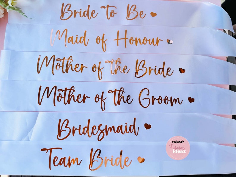 Peut inclure: Six rubans de satin blanc avec des lettres dor&eacute;es roses. Les rubans indiquent "Bride To Be", "Maid of Honour", "Mother of the Bride", "Mother of the Groom", "Bridesmaid" et "Team Bride".