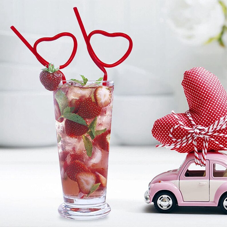 May include: Two red heart-shaped straws in a glass of strawberry lemonade with ice and mint leaves. A pink toy car with a red heart-shaped pillow on top.