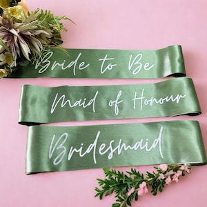 May include: Three sage green satin sashes with white script: "Bride to Be", "Maid of Honour", and "Bridesmaid". The sashes are arranged on a pink surface, with floral accents at the top and bottom.