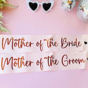 Peut inclure: Deux sashes en satin rose avec des lettres dor&eacute;es. La sangle sup&eacute;rieure indique "M&egrave;re de la mari&eacute;e" avec un petit c&oelig;ur dor&eacute;. La sangle inf&eacute;rieure indique "M&egrave;re du mari&eacute;" avec un petit c&oelig;ur dor&eacute;.
